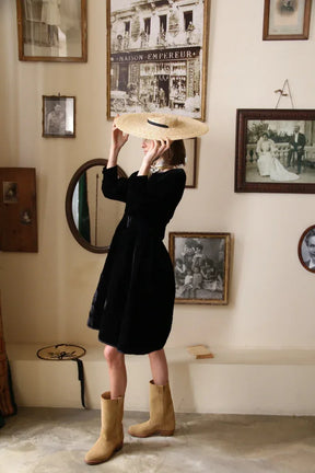 Woman in a black dress with tan boots holding a wide-brim straw hat, standing by vintage framed photos.