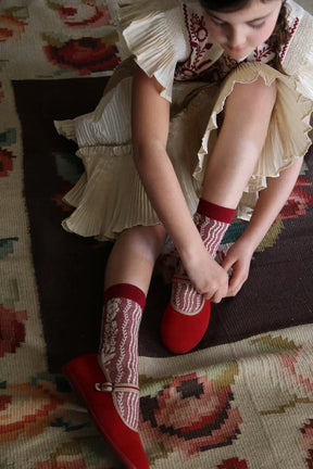 Girl in pleated cream dress with red embroidery, wearing red socks and shoes on floral rug