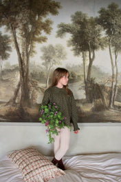 Girl in patterned green blouse holding flowers stands on bed, vintage forest mural background