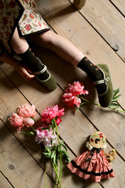 Child in floral dress and green Mary Jane shoes on wood floor with pink peonies and fabric doll