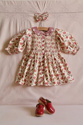 Floral smocked baby dress with peter pan collar, matching hair bow, and red clogs on pink linen