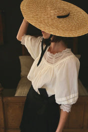 Woman in a vintage white lace blouse and wide straw hat, standing indoors