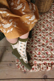 Child in yellow embroidered dress, floral socks, and green Mary Jane shoes on patterned rug