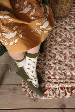 Child in yellow embroidered dress, floral socks, and green Mary Jane shoes on patterned rug