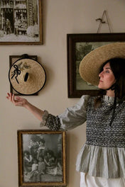 Woman in a wide-brimmed straw hat and patterned blouse standing by vintage framed art and a hanging straw hat