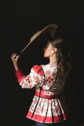 Woman in floral quilted jacket with red accents holding straw hat against dark background