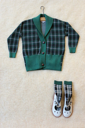 Green plaid children's cardigan with matching socks and white shoes on beige fabric