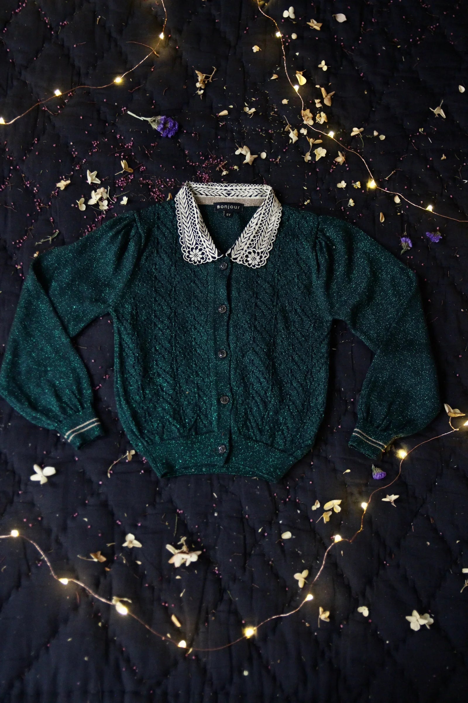 Green knit children's cardigan with white lace collar, displayed on dark fabric with fairy lights and flower petals.